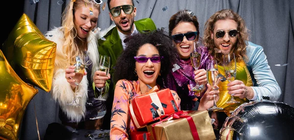 Sonrientes amigos interracial con globos bebiendo champán cerca de cortina gris sobre fondo negro, pancarta - foto de stock