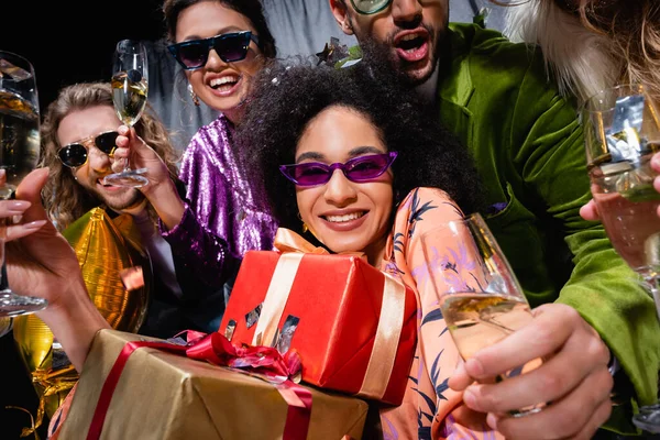 African american woman in sunglasses holding gift boxes among interracial friends near grey curtain on black background — Stock Photo