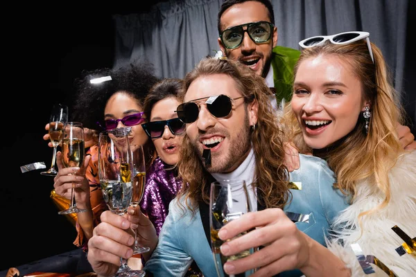 Glückliche interrassische Freunde sitzen mit Champagner in der Nähe des grauen Vorhangs auf schwarzem Hintergrund — Stockfoto