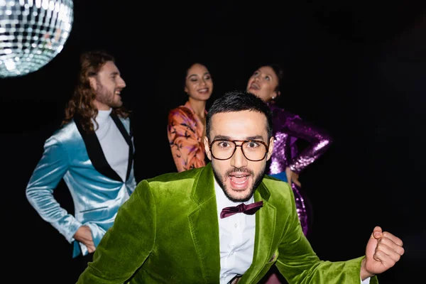 Позитивний арабський чоловік в окулярах дивиться на камеру на вечірці з міжрасовими друзями на чорному тлі — стокове фото