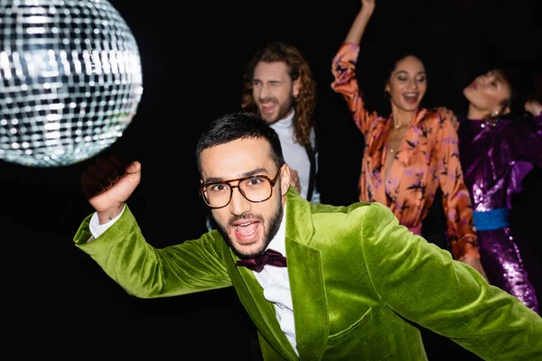 Positif jeune homme arabe adulte dans des lunettes de danse avec des amis multiraciaux en vêtements colorés dans une boîte de nuit sur fond noir — Photo de stock
