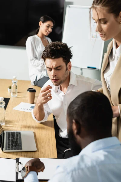 Hombre de negocios reflexivo cerca de la computadora portátil y socios comerciales multiétnicos, borrosa primer plano - foto de stock