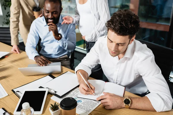 Uomo d'affari che scrive nel quaderno vicino ai colleghi multietnici nella sala conferenze — Foto stock