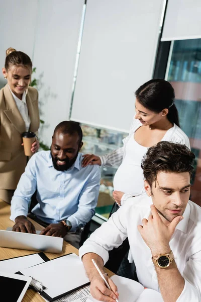 Sonriente hombre de negocios afroamericano escribiendo en el portátil cerca de colegas multiétnicos - foto de stock