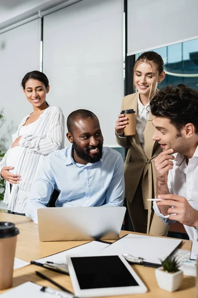 Hombre de negocios afroamericano sonriendo cerca del ordenador portátil y colegas positivos durante la reunión - foto de stock