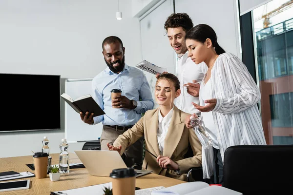 Sorridente empresária apontando para laptop perto de felizes colegas inter-raciais na sala de conferências — Fotografia de Stock