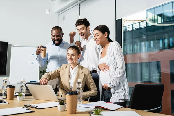 Smiling businesswoman pointing at laptop near excited interracial colleagues — Stock Photo