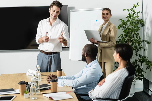 Sonriente hombre de negocios mostrando pulgares hacia arriba durante la reunión con colegas multiétnicos - foto de stock