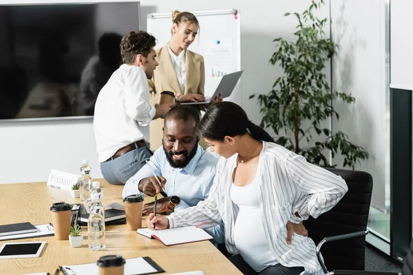 Smiling african american man pointing at notebook near pregnant businesswoman and talking colleagues — Stock Photo