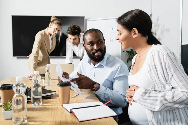 Усміхнений афроамериканський бізнесмен вказує на документ біля вагітної колеги — стокове фото