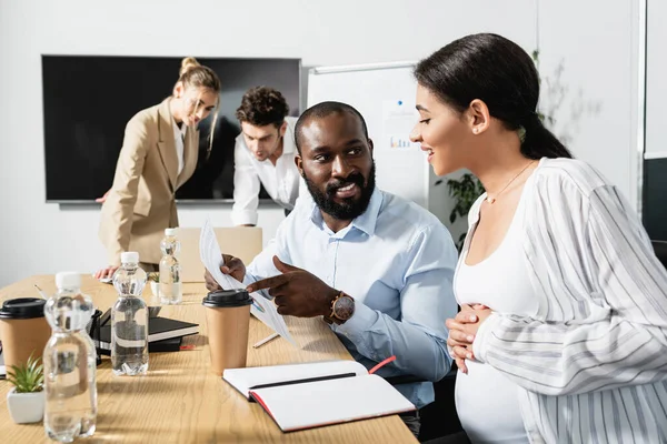 Sonriente hombre de negocios afroamericano apuntando a documento cerca de colega embarazada - foto de stock