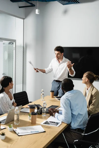 Joven empresario sosteniendo periódico mientras habla con compañeros de trabajo multiétnicos - foto de stock