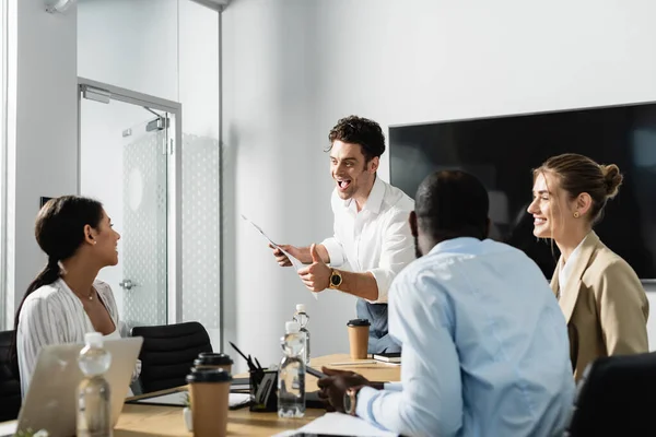 Aufgeregter Geschäftsmann mit Zeitung zeigt Daumen hoch bei multiethnischen Kollegen — Stockfoto