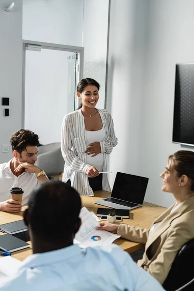 Donna d'affari africana americana incinta sorridente mentre indica il computer portatile vicino ai partner commerciali — Foto stock