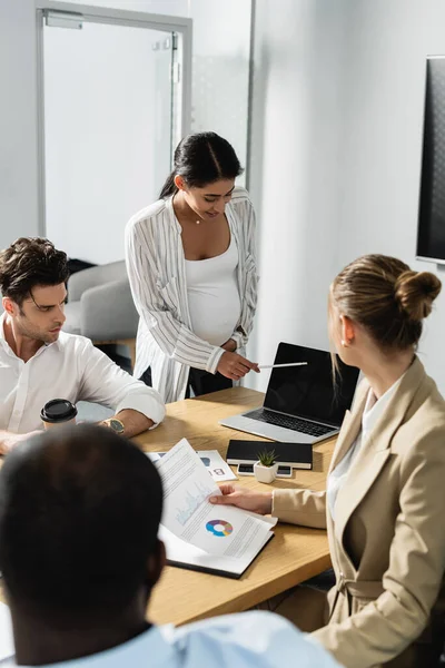 Donna d'affari africana americana incinta che punta al computer portatile vicino a partner commerciali multiculturali — Foto stock