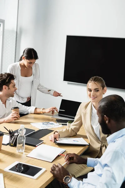 Joven empresario apuntando a la computadora portátil con pantalla en blanco cerca de colegas multiétnicos - foto de stock