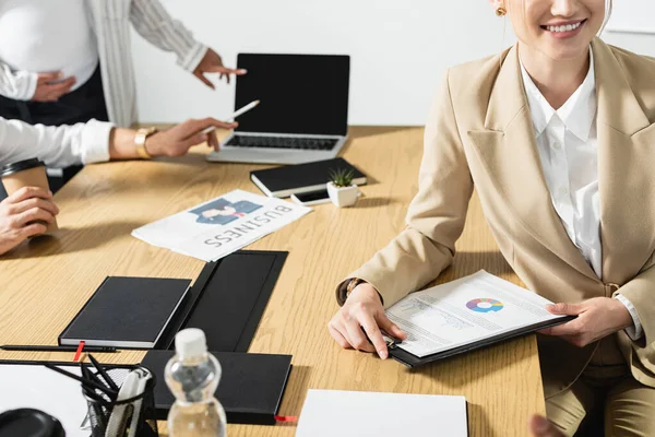 Partial view of young businesswoman smiling near infographics and blurred interracial colleagues — Stock Photo