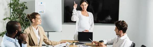 Pregnant african american businesswoman pointing with finger while talking to business partners, banner — Stock Photo