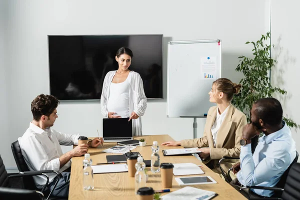 Pregnant african american businesswoman pointing at laptop during discussion with multiethnic coworkers — Stock Photo