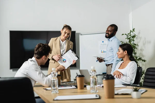 Smiling businesswoman pointing at graphs while talking to multiethnic coworkers — Stock Photo