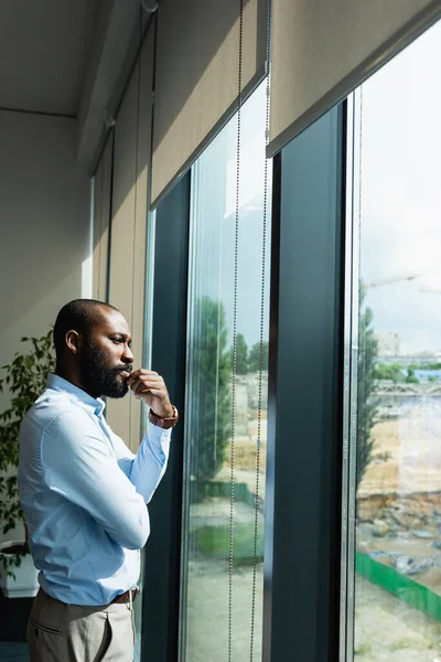 Thoughtful african american businessman looking away while standing near window — Stock Photo