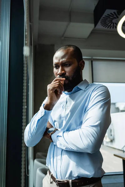 Pensativo hombre de negocios afroamericano cogido de la mano cerca de la cara mientras está de pie en el cargo - foto de stock