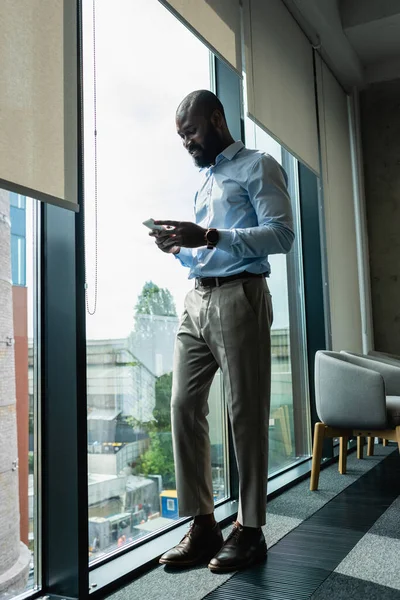 Sorridente uomo d'affari africano americano messaggistica su smartphone vicino alla finestra — Foto stock