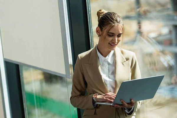 Lächelnde Frau benutzt Laptop in der Nähe von Fenster im Büro — Stockfoto