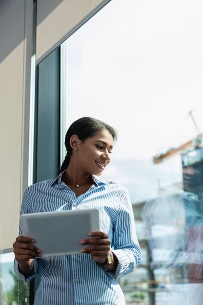 Feliz mulher de negócios afro-americana olhando através da janela enquanto segurando tablet digital — Fotografia de Stock