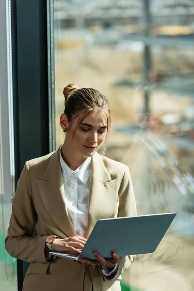 Young businesswoman standing near window in office with laptop — Stock Photo