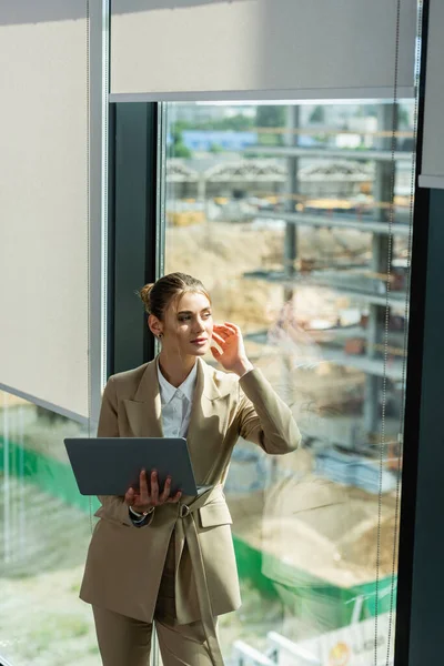 Mujer de negocios soñadora sosteniendo el ordenador portátil mientras mira a través de la ventana en la oficina - foto de stock