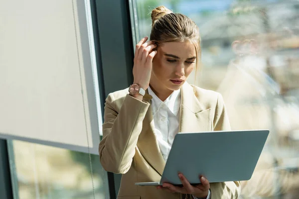 Вдумчивая деловая женщина чинит волосы, глядя на ноутбук в офисе — стоковое фото