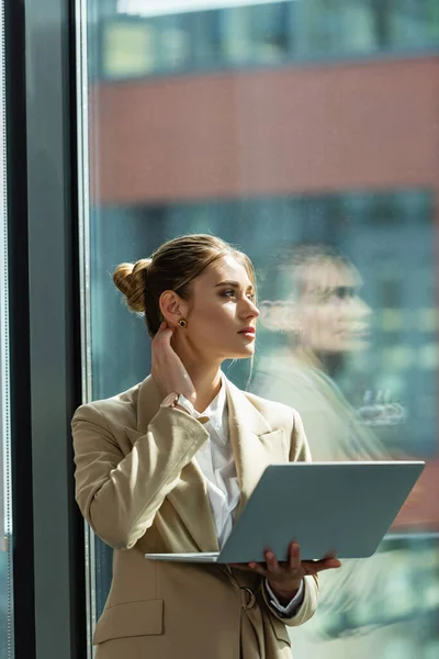 Joven mujer de negocios con portátil pensando cerca de la ventana en la oficina - foto de stock