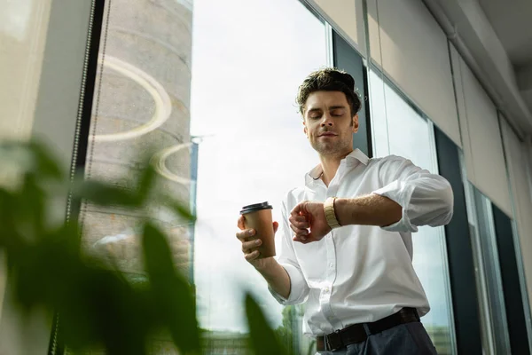 Homme d'affaires avec café pour aller regarder la montre-bracelet près de la fenêtre sur le premier plan flou — Photo de stock