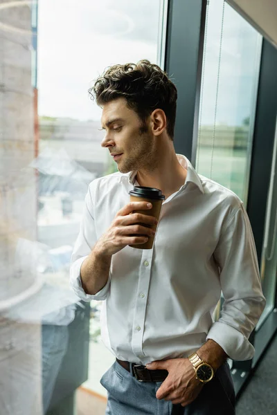 Jeune homme d'affaires avec tasse en papier regardant par la fenêtre dans le bureau — Photo de stock