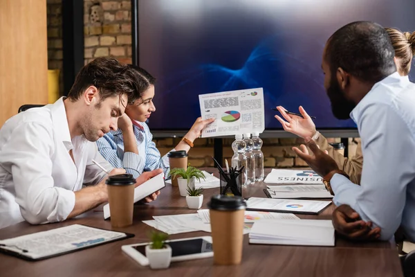 African american businesswoman holding document with analytics near multiethnic colleagues — Stock Photo