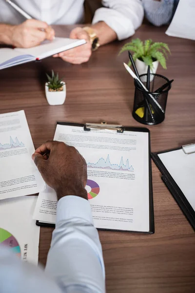 Vista recortada de hombre de negocios afroamericano cerca borrosa colega escribir en cuaderno - foto de stock