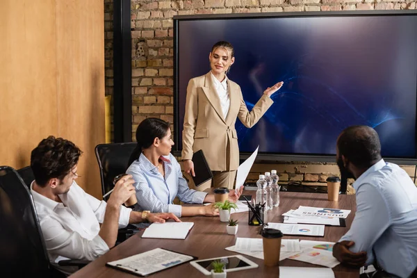 Businesswoman pointing at lcd monitor near multiethnic colleagues during meeting — Stock Photo