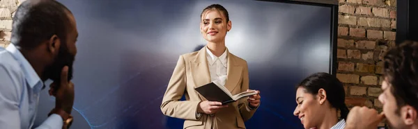 Femme d'affaires souriante debout avec ordinateur portable près de collègues interraciaux, bannière — Photo de stock