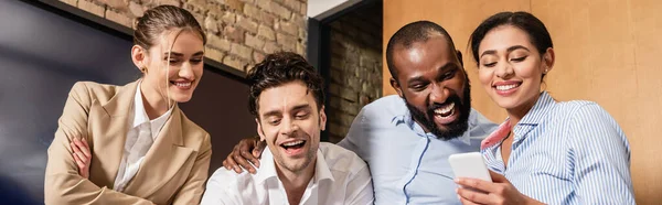 Mujer afroamericana feliz sosteniendo teléfono inteligente cerca de compañeros de trabajo alegres, pancarta - foto de stock