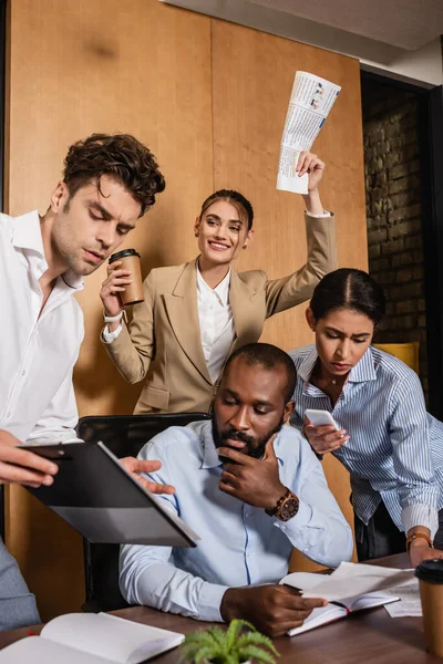 Excited businesswoman holding newspaper near thoughtful interracial colleagues — Stock Photo