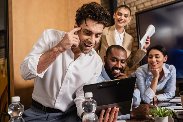 Aufgeregter Geschäftsmann zeigt Ideengeste beim Blick auf Klemmbrett neben lächelnden multiethnischen Kollegen — Stockfoto