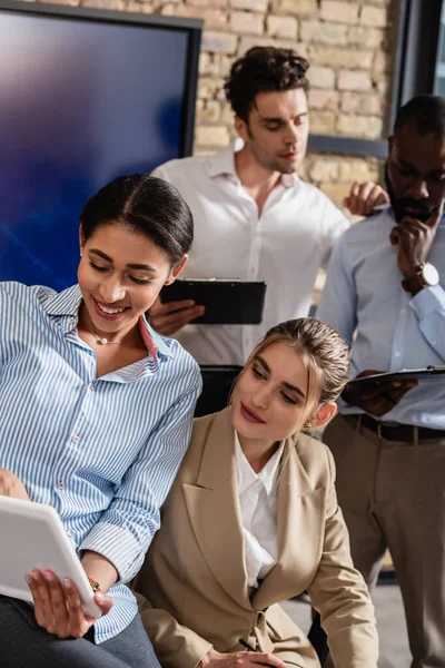 Smiling interracial businesswomen looking at digital tablet near blurred colleagues — Stock Photo