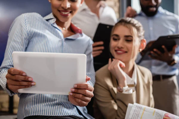 Selective focus of digital tablet in hands of smiling african american businesswoman near blurred colleagues — Stock Photo