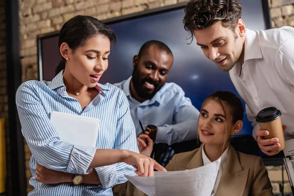 Empresaria afroamericana señalando documentos cerca de compañeros de trabajo sonrientes - foto de stock