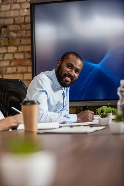 Bem sucedido empresário afro-americano sorrindo para a câmera perto do monitor lcd na sala de conferências — Fotografia de Stock