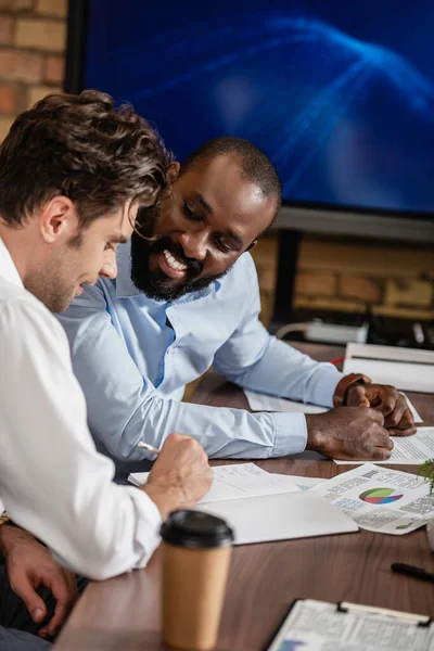 African american businessman smiling near colleague writing in notebook during meeting — Stock Photo