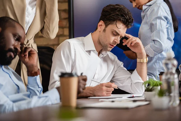 Thoughtful businessman working near interracial colleagues on blurred foreground — Stock Photo