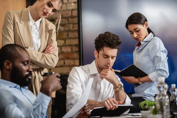 Thoughtful businessman looking at notebook near multiethnic colleagues — Stock Photo