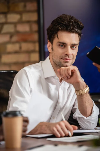 Homme d'affaires souriant regardant la caméra alors qu'il était assis au bureau, flou au premier plan — Photo de stock